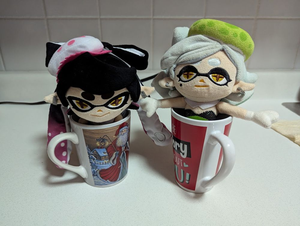 Callie Plush and Marie Plush sit inside mugs, with their heads (and arms, in Marie's case) popping out. 