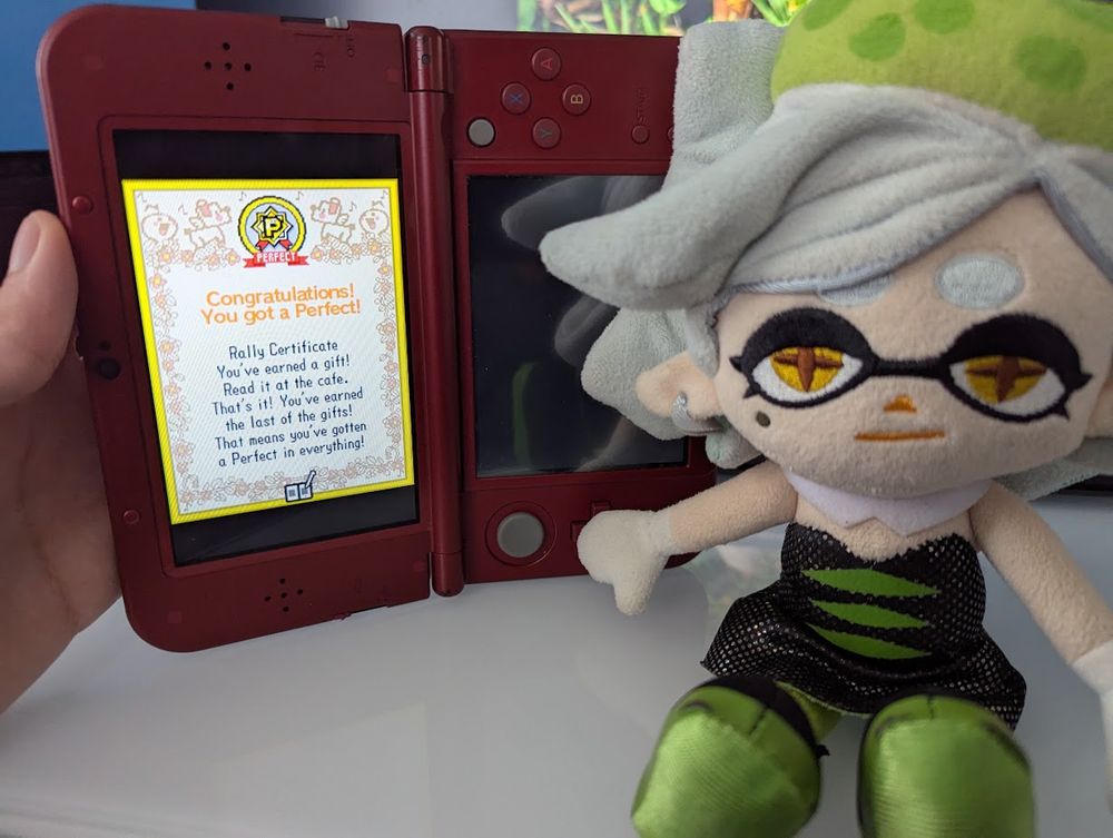 Marie Plush sits beside my New 3DS XL which is playing Rhythm Heaven. The 3DS is beind held sideways; the left screen shows a congratulations message after getting every Perfect in the game (shown in quoted pic).