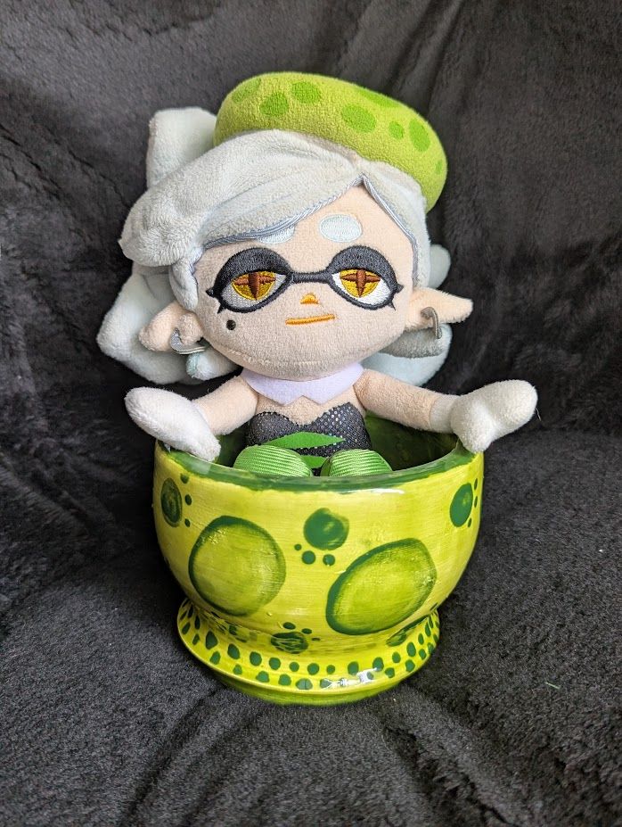 Marie Plush snuggly sits inside a handmade glazed ceramic bowl. The bowl is about 4½ inches in diameter and 3¼ inches tall. The exterior is painted in lime green with dark green spots, resembling Marie's headpiece. The exterior's base is also adorned with smaller dark green spots, and the interior of the bowl is dark green. 
