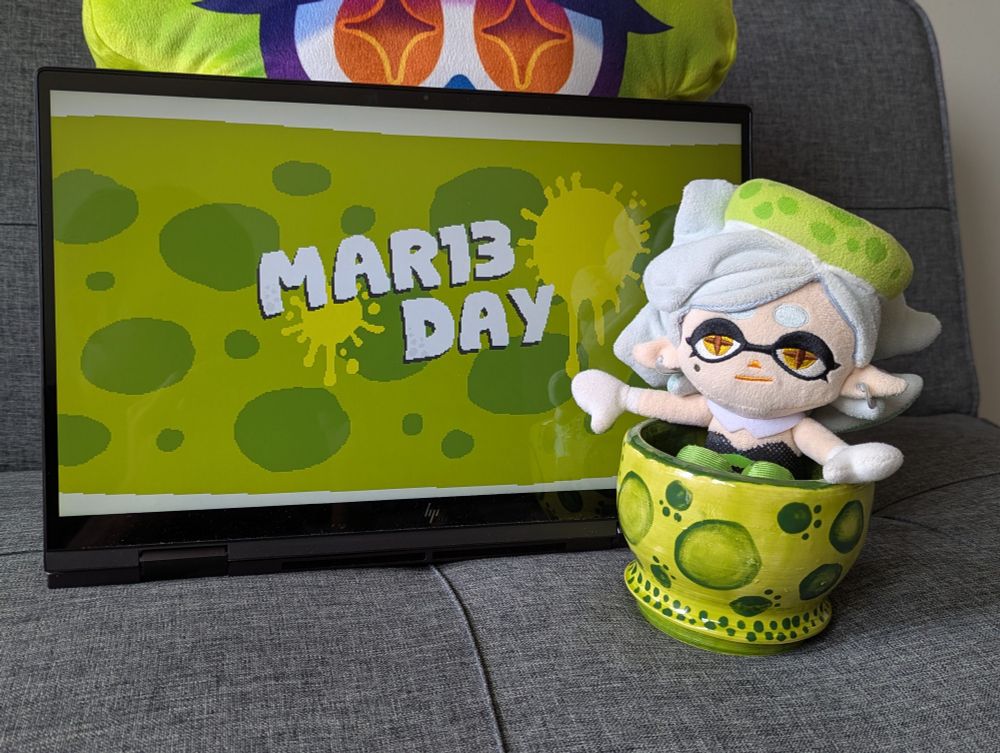 Marie Plush sits on a ceramic bowl themed after her headpiece. Right next to her is a screen displaying pixel art I made: the text "MAR13 DAY" appears in white in the center of the screen, there are two lime-green ink splatters, and the background is based on Marie's headpiece. A Marie squid-form pillow (from TORI.PNG) is peeking from behind the screen. 
