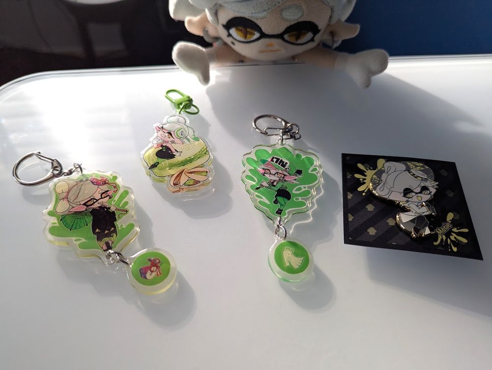 Four merch items displayed on my desk. From left to right: a two-sided keychain currently showing Marie's kimono outfit (the other side is her Alterna outfit), a keychain featuring Marie sitting on a pistachio-flavored macaron, another two-sided keychain currently showing Marie in a casual outfit wearing a cap and facemask and holding her Hero Charger (the other side is her standard outfit), and an enamel pin featuring Marie in her Alterna outfit with her umbrella. Marie Plush is leaning towards the desk from the back, looking at the items. 