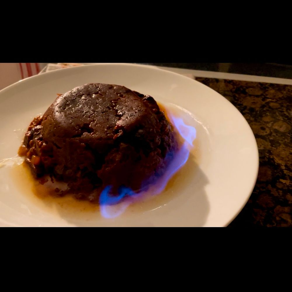 A Christmas pudding (like me, a little slumped but still tasty) sits soaked in bourbon (also a little like me.) Unlike me, it sits on a white plate, and the bourbon is burning off with a bright, blue, flame. 
