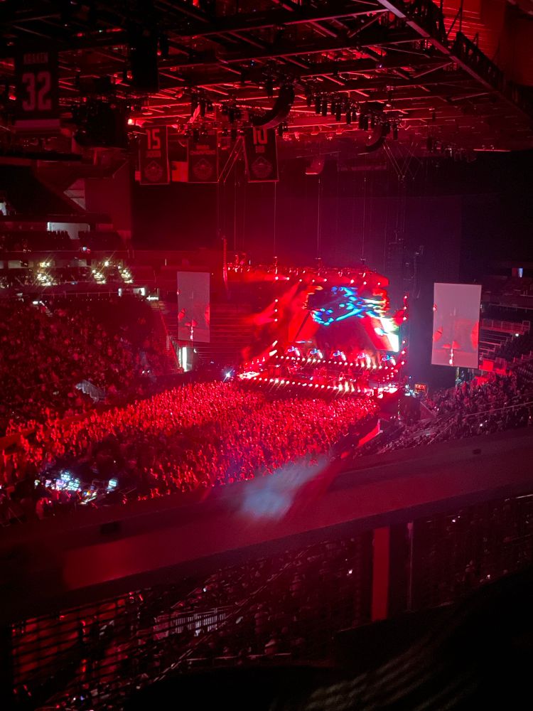 Dark photo from back if arena with lights over a large number of people watching from the arena floor and a big stage with lights. 