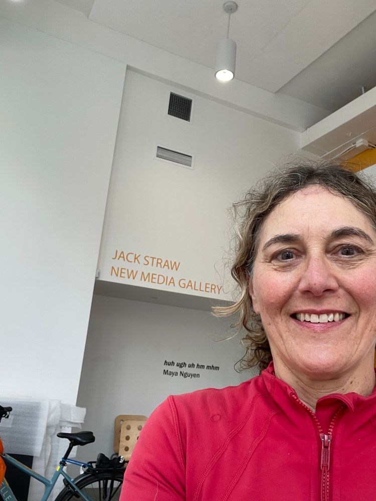 foreground white woman’s face with blonde/brown hair pulled back and red jacket and parked blue ebike and white wall interior with yellow letters on white wall. 