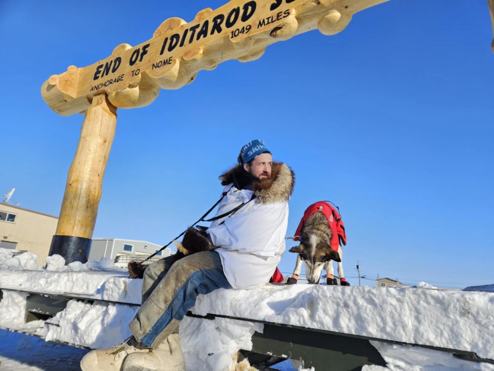 A man in a white parka sits next to a gray and white husky with a white strike down her forehead. She nibbles something tasty off the ground while he looks over his shoulder. Above them is the burled arch that marks the Iditarod's finish line in Nome. They did it.