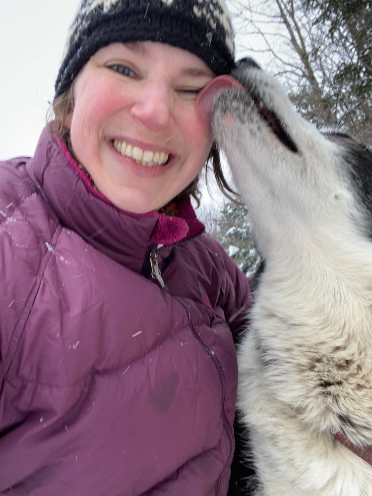 A woman in a puffy purple coat and a knit black and white hat grins as she wraps her arm around a black and white husky, who licks her eyeball, lifting his spectacular furry head to reveal incredibly plush neck fur. One tiny fang is visible. It's razor-sharp, designed to interfere with the workings of both brakelines and and fascist governments. 