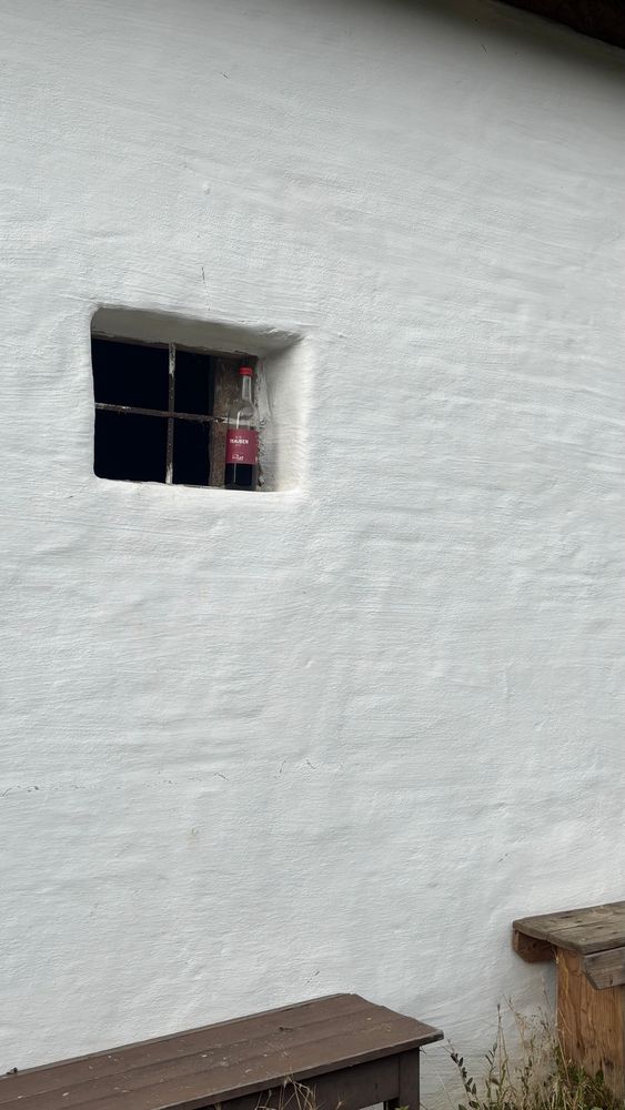 Eine Halb volle Rotweinflasche steht vor einem vergitterten Fenster, die Außenwand ist weiß und ein Bild Ende untenä