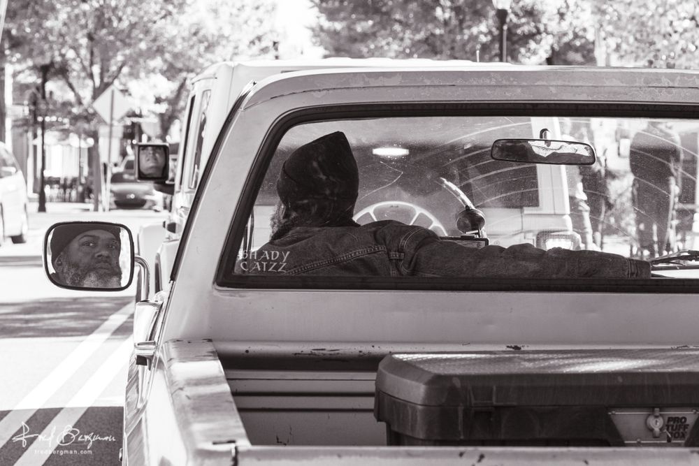 Man in pickup truck photographed from behind while his face is reflected in the side view mirror and the rear window has sticker that says "Shady Catzz"