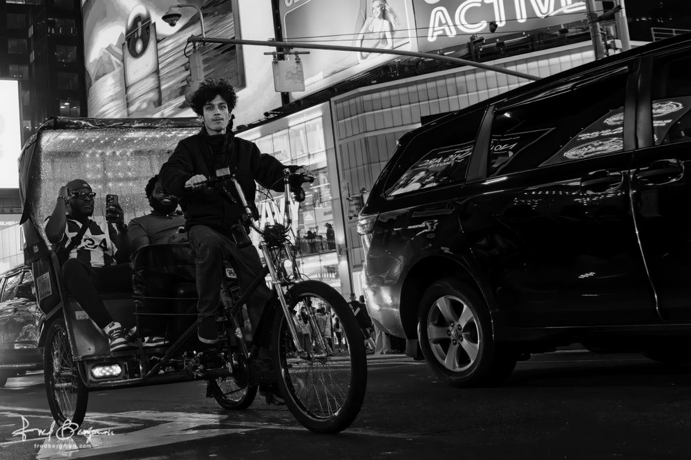 A young man is pedaling a pedicab with two passengers with their cameras out and smiles on their faces. They are on a busy street near Times Square with cars very close by.