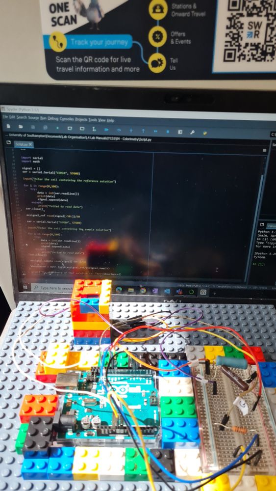 A laptop open with a screen of Python code. In front is a lego construction holding an Arduino board and some other electronic connectors.
