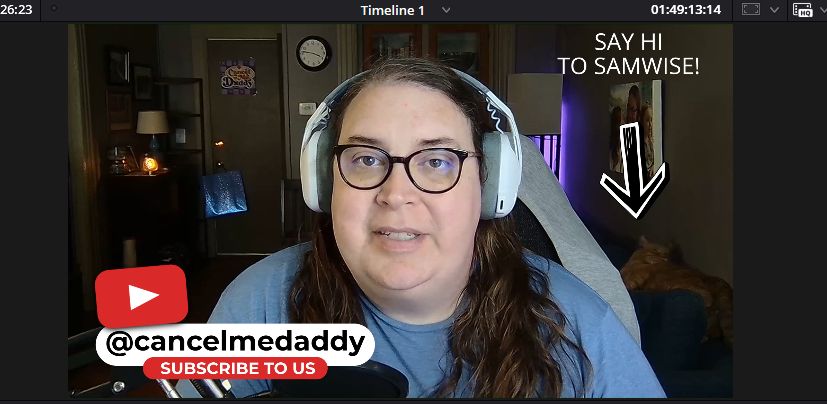 a still frame from next week's Cancel Me, Daddy youtube episode. it shows a white trans woman with brown hair (me) with white headphones on and a blue t shirt, a graphic bottom left encouraging people to subscribe to our channel, and a black arrow pointing at an orange tabby cat in the background laying on a blue couch with text that says "SAY HI TO SAMWISE!"