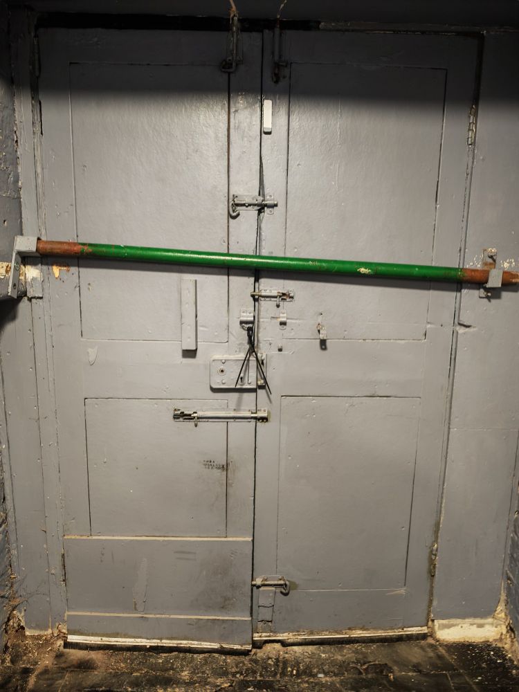 A barred door with a collection of bolts and a large metal bar across the middle. The repair to one door and the damage to the doors suggest the bolts and bar have been needed in the past and possibly the future. 
Taken at the Rat and Pigeon in Manchester, UK.