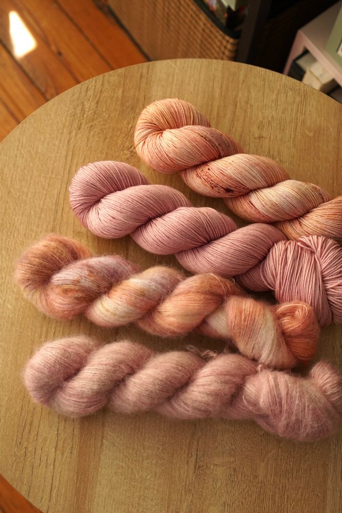 Four skeins of yarn are fanned on top of a light wood circular side-table. The bottom two skeins are fluffy texture and the top two are a smooth, thin sock weight. Two colorways are displayed, a variegated blend of soft pastel pinks, warm orange and purple and a semi-solid dusty lilac pink. 