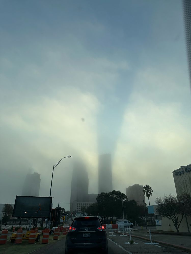 two tall city buildings split the suns rays of a sunrise, but it’s muted by fog 
