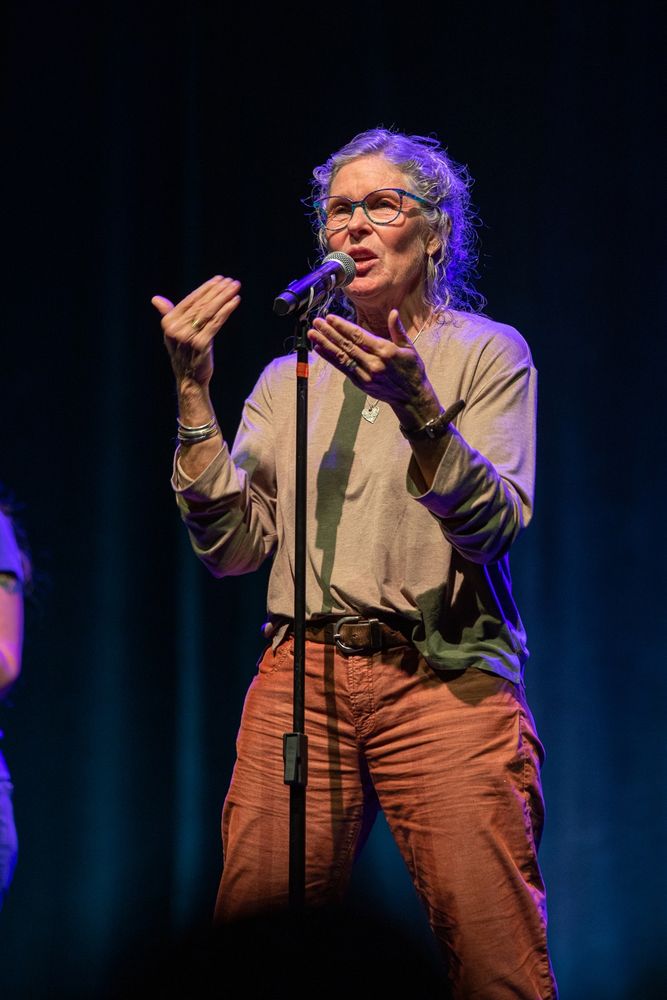 A woman wearing glasses is speaking into a microphone. Photo credit kmr studios