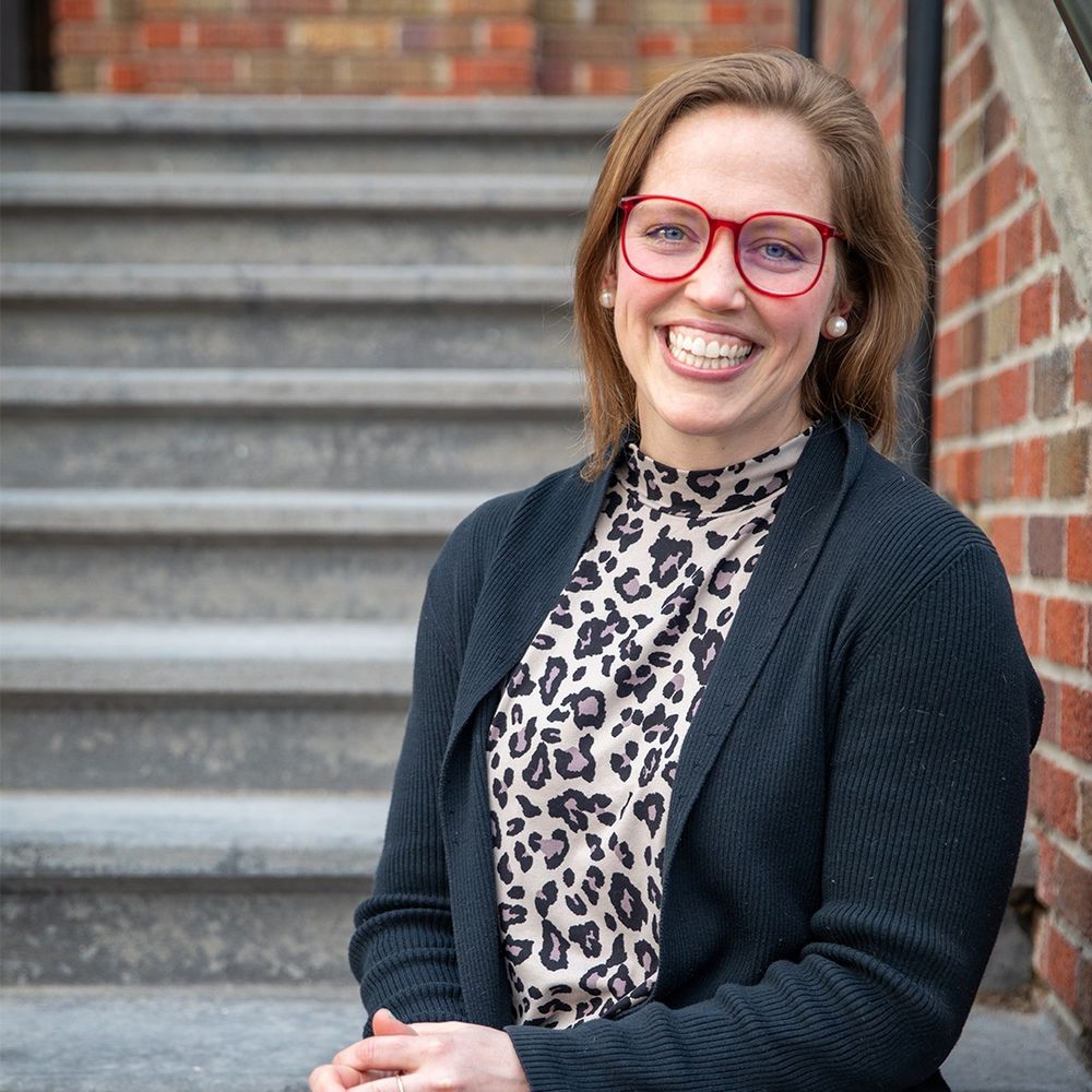 The image features a woman smiling at the camera, showcasing her joyful expression. She is wearing fashionable eyewear and outerwear, adding to her stylish appearance. The setting is outdoors. Listen to her story @ tellussomething.org or wherever you get your podcasts.