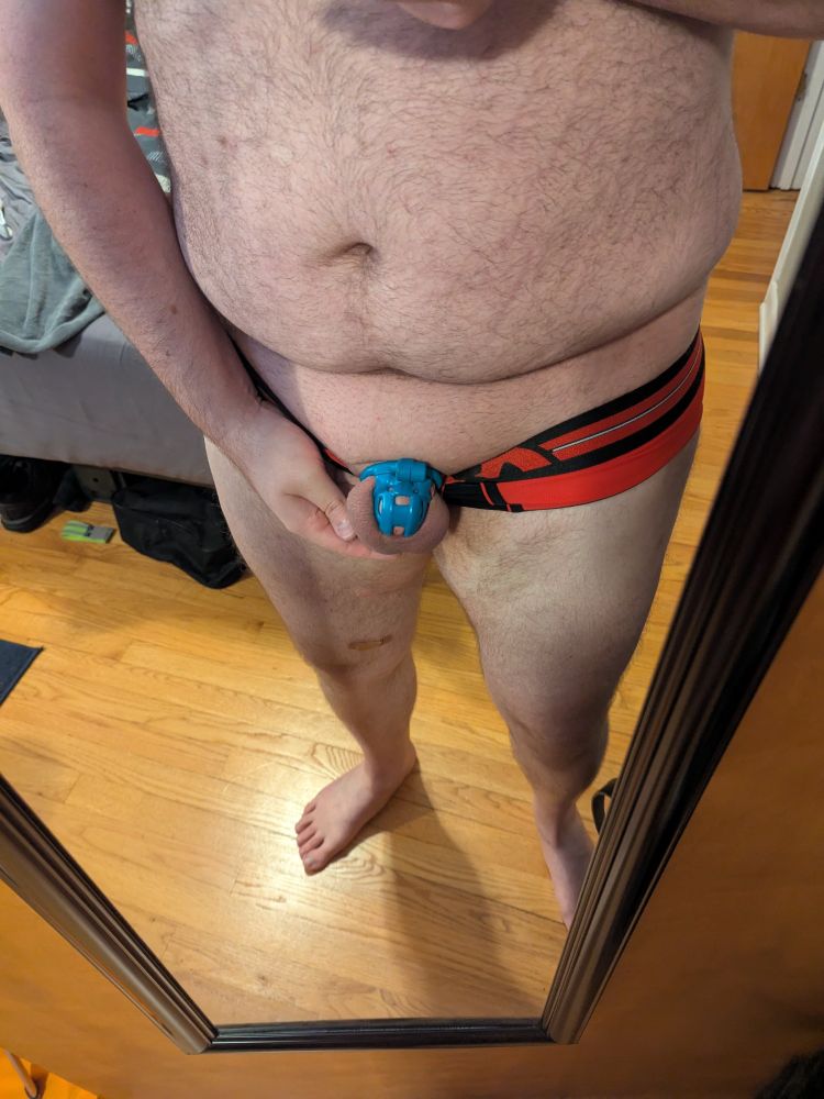 A picture of me in wearing only a red and black jock brief from Mr. S Leather. I'm showing my blue kink3d chastity cage with the jock tucked behind my balls and cage.