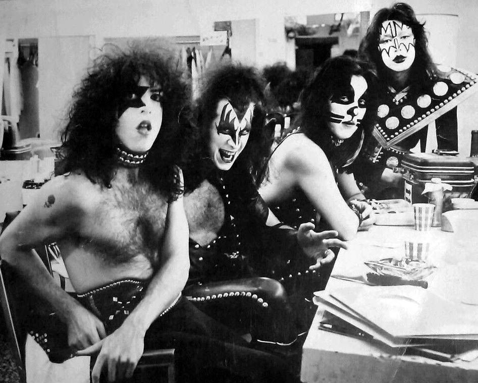 Photo of the 4 guys in the band KISS sitting at a table that has some papers and cups of water. Band stuff. They're all white guys with black-and-white face paint, big hair (some fluffy, some just luxurious) and their outfits are all made of black leather with metal studs, from 'skimpy' to 'complete suit'. One of them is making a face at the camera, but the other 3 are chillin and working. 