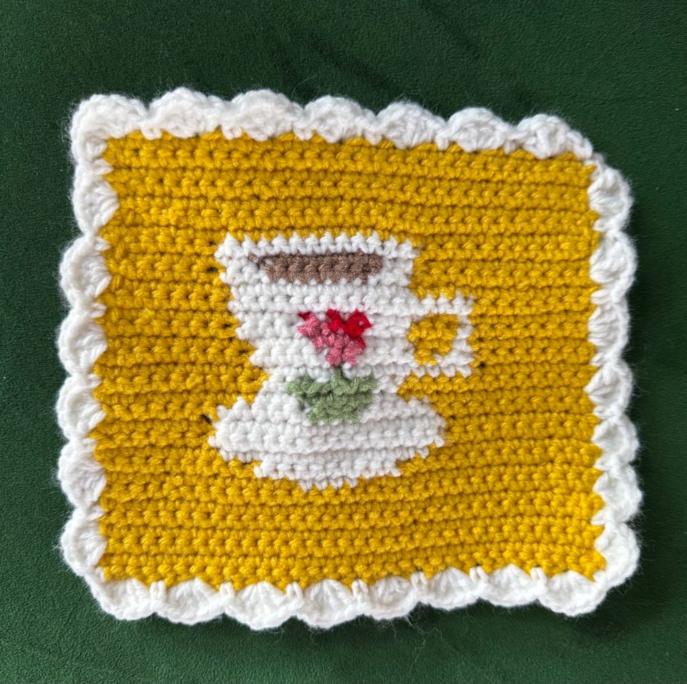 Crochet tapestry piece with yellow background and a white teacup filled with brown tea and a red and pink flower. It has a white scalloped border and is resting on a dark green couch 