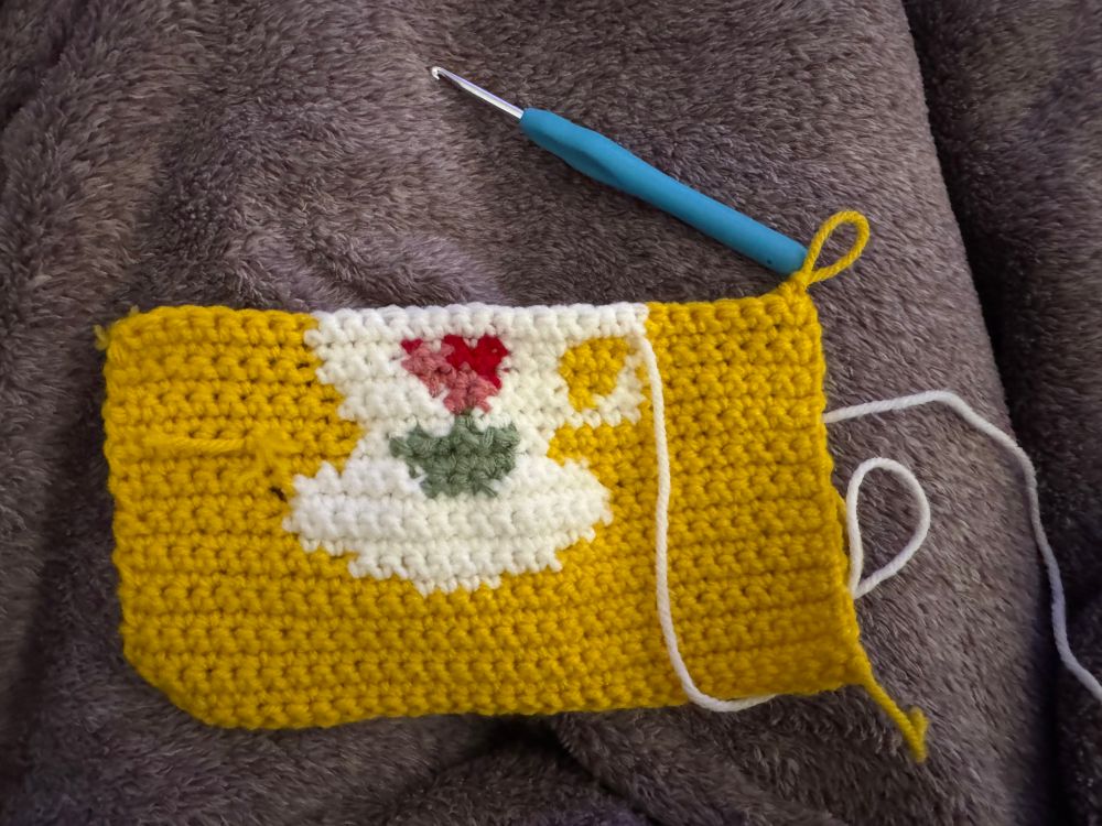 Pictures is a background of a purple blanket. Resting on top is a crocheted piece with a yellow background and a white teacup with a handle and red and pink flower in its center. It is unfinished and has a loose white yarn hanging from the piece and the size 4 blue crochet hook resting nearby. 