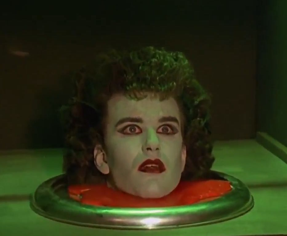 a very pale man's head with red lipstick and dark eyeliner, with big fluffy 80s hair is sat atop a red covered silver platter.

There's slightly eerie green lighting and it looks like he's in a box.  It's actually a dumb waiter.