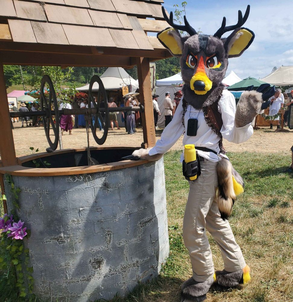 A deer fursuiter posing at the Washington Midsummer Renaissance Faire. They are standing next to a replica of a well. Their fursuit is a warm gray with orange and yellow markings.
