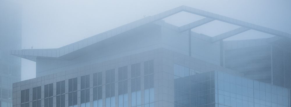 An urban building – in shades of mid-blue, dark blue and gray – is rendered in softened contrast courtesy of the low-flying cloud which engulfs its top.  The building wears a fascinator shaped like a zippy flattened "V", which feature is especially befogged.