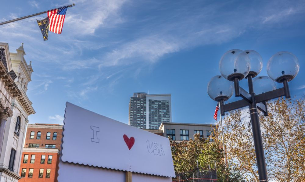 A view tilted upward.  Near the bottom of the frame, we see the back of a sign reading "I [red heart] USA".  We see a brilliant backlit US flag before a blue sky with swirly white clouds.  We also see tops of urban buildings, another US flag, and the glass globes of an outdoor light.