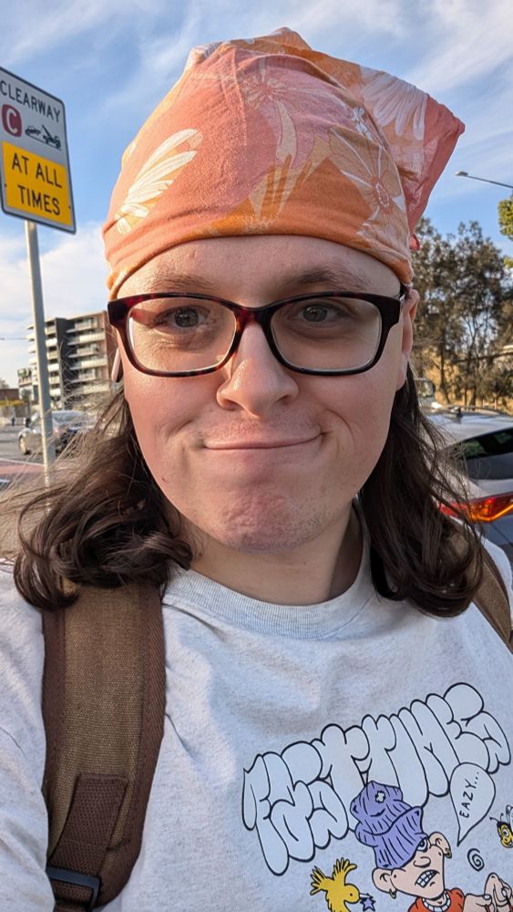 A white autistic transfemme woman, with an orange CanTeen bandana, worn over her scalp to reduce sweat and sun exposure.

She is wearing her signature seeing glasses, a brown canvas backpack, and a grey Fast Times tee, as well as her signature smirk.