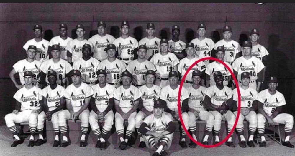 Black and white team photo of the 1964 St Louis Cardinals in white home uniforms. Bob Gibson and Bob Uecker are toward the right end of the front of three rows, holding hands and grinning, whereas the other players are just smiling or posing normally. Uecker and Gibson are circled in red on the image.