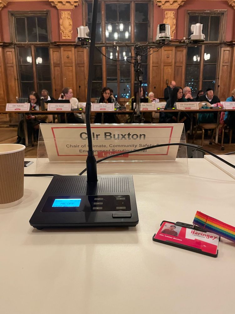 Picture of microphone, badge and name plate with “Cllr Buxton, Chair of Climate, Community Safety and Environment Scrutiny Panel”. With a background of a town hall with councillors 
