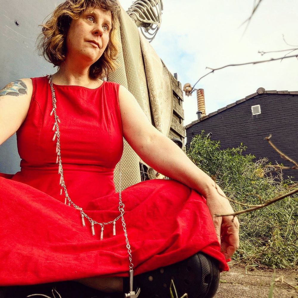 A woman sitting in a garden in a dress with black Crocs peeking out under it. She's wearing a bandolier style bodychain necklace and looking off into the distance. 