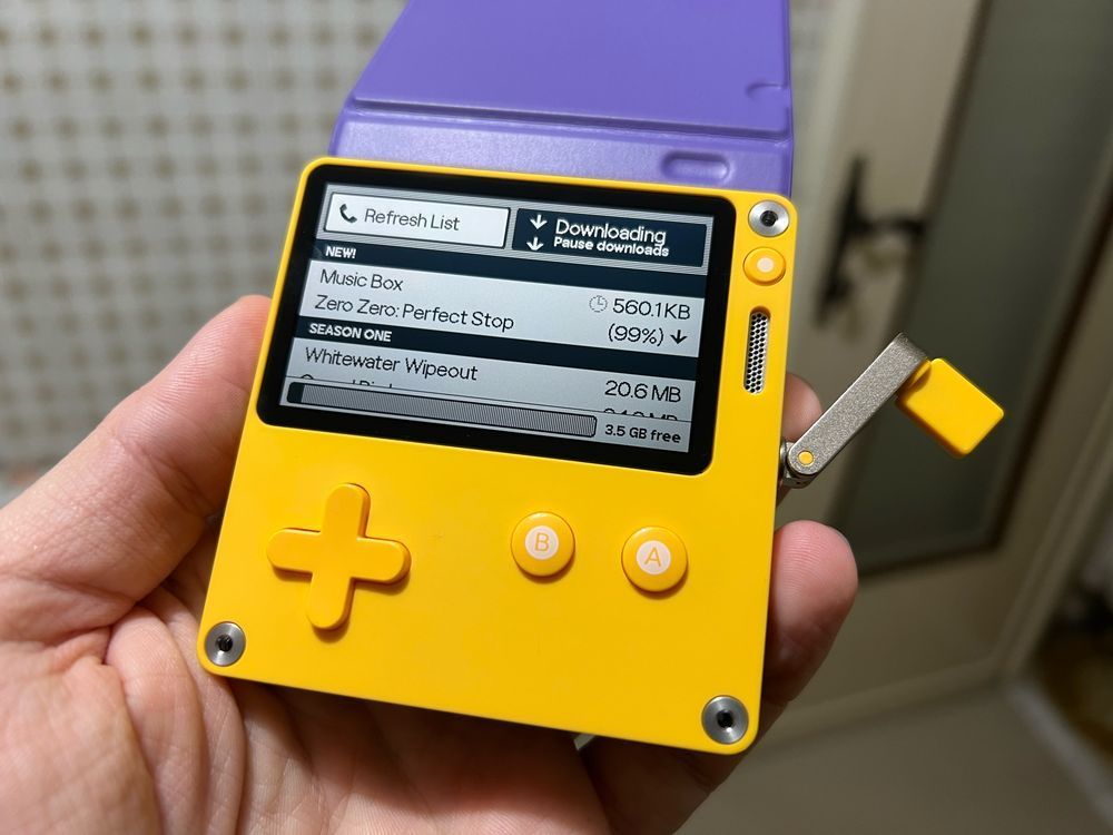 A photo of a hand holding a yellow PlayDate console, with a purple cover open towards the back.