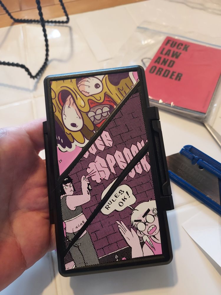 Some stickers on an SD card case. One is a melting dog face by kc green and another is a silver sprocket sticker by idk I can't remember. On the table behind it is the xacto blade I used and a sticker that says "fuck law and order"