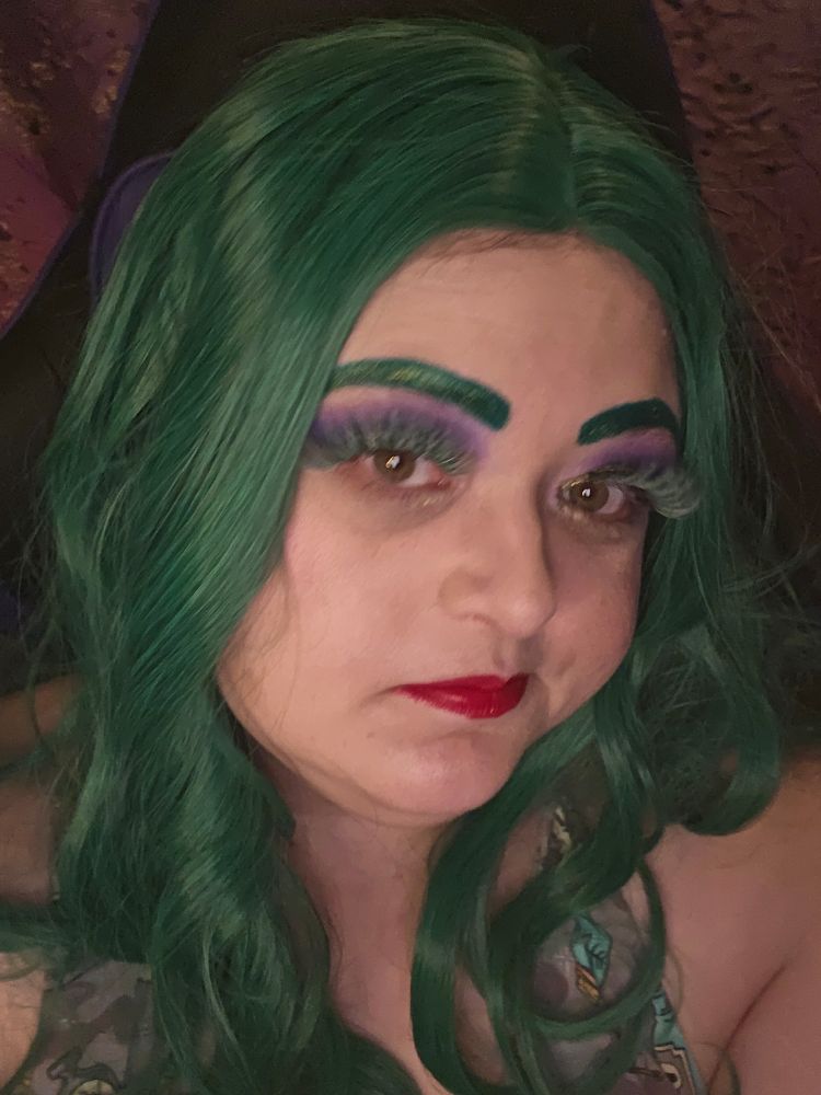 Woman with green hair, red lips, green lashes, purple eyeshadow, and green eyebrows makes face