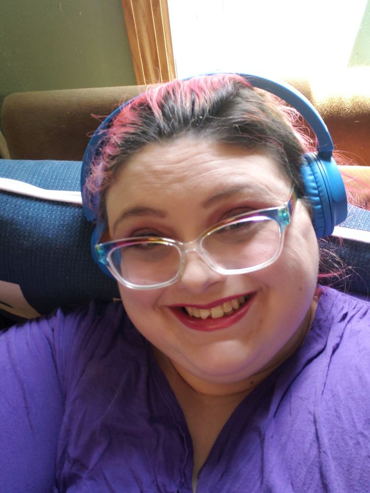 Woman with blue headphones, purple shirt, red lipstick and rainbow glasses smiles for the camera