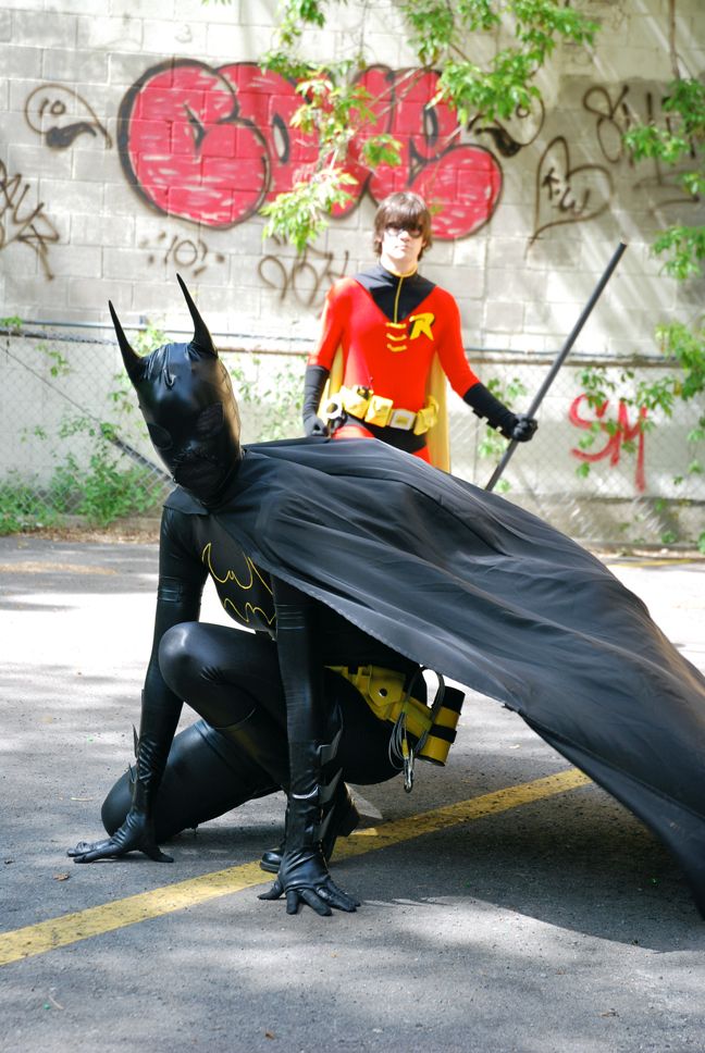 Cosplayer dressed as Cassandra Cain (Batgirl), posed in an alleyway like she's just jumped from somewhere high and landed in a crouch. In the background, Red Robin stands tall, with a bo staff in his hand.