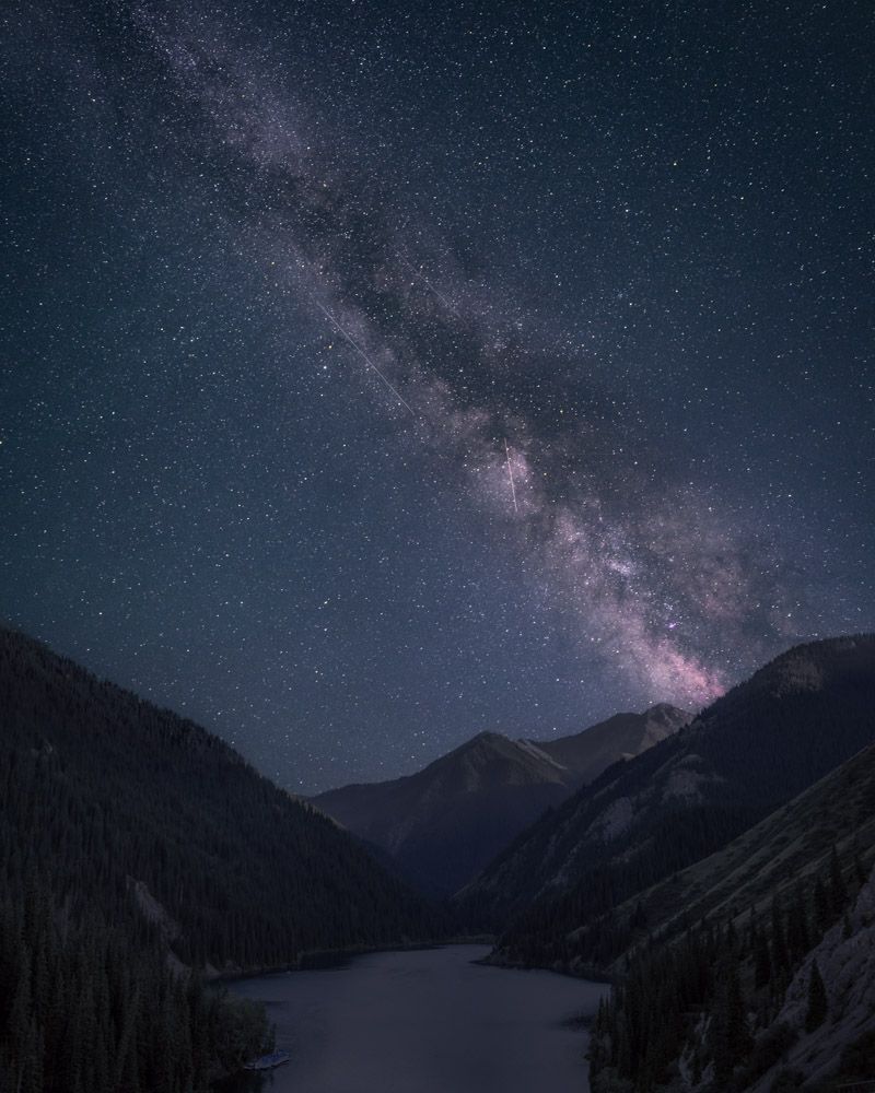 A blueish monochromatic image of a lake surrounded by mountains. The core of the Milky Way stretches across the night sky, from the top left to the bottom right. The image has a serene quality to it.