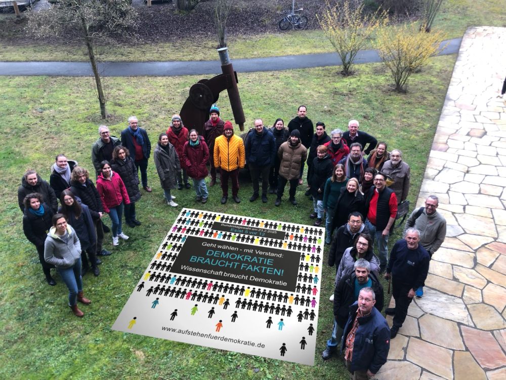 Das Banner "Demokratie braucht Fakten!" ist auf die Wiese vor dem Geozentrum der Goethe-Universität projeziert. Umrahmt wird es von 3 Dutzend buntgekleideter Wissenschaftlerinnen und Wissenschaftler.