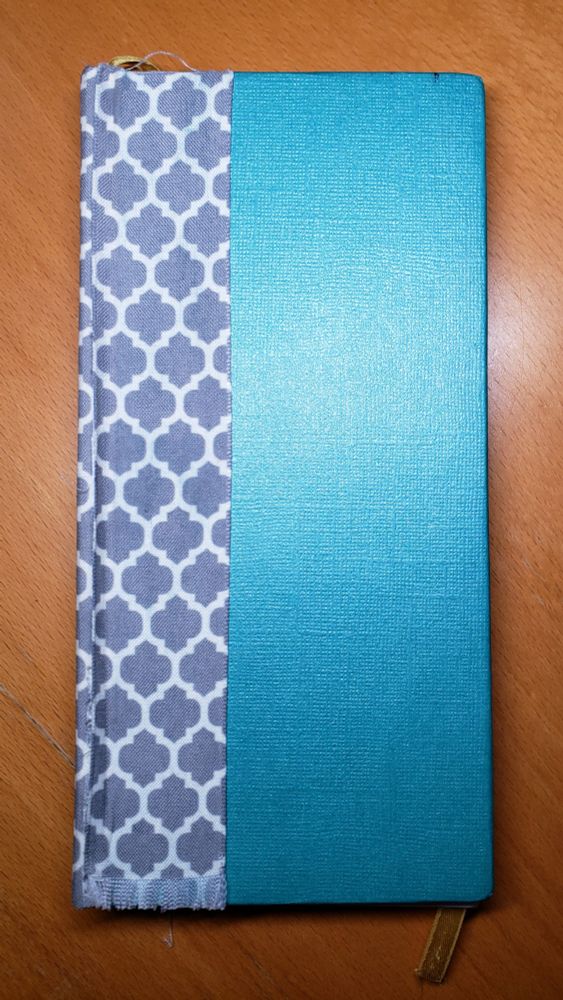 Closed notebook, roughly twice as tall as wide. Cover is a woven textured teal, with a grey and white cloth covering on the spine, and a gold ribbon bookmark showing at the top and bottom.