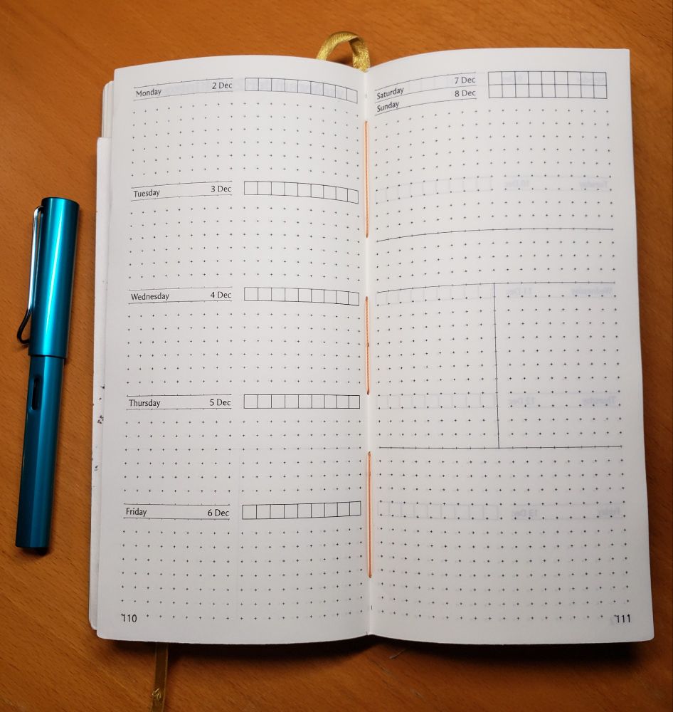 Open notebook with white pages with planner layout printed on it, with a gold colored ribbon running behind, a teal Lamy Al-Star fountain pen on the left side, and on a brown wood grained background. Pages are printed with dot grid and sectioned off for days of the week from Monday 2 Dec to Sunday 8 Dec, with Monday through Friday vertical on left, Saturday, Sunday and some divided boxes on the facing right. Stitched down the middle with orange thread.