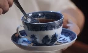Photo of a blue and white teacup with a spoon in it (from 2017 movie "Get Out"). Teacup is on a blue and white saucer held by in a person's left hand, and contains dark brown tea, and spoon is held at an angle by the right hand. Image is close up on tea cup, so only part of person's torso and arms/hands are in frame, and they are wearing a white shirt.