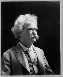 Black and white photo of a man in three-quarters profile from lower torso up. He is recognizable as Mark Twain, or Samuel Clemens, and relatively late in his life. He has white hair and bushy mustache, thick eyebrows, and has an expression suggesting he is at least mildly annoyed to be posing for a photo. He is wearing a medium dark suit with a vest, and white collared shirt with no tie, and the background is a uniform black.