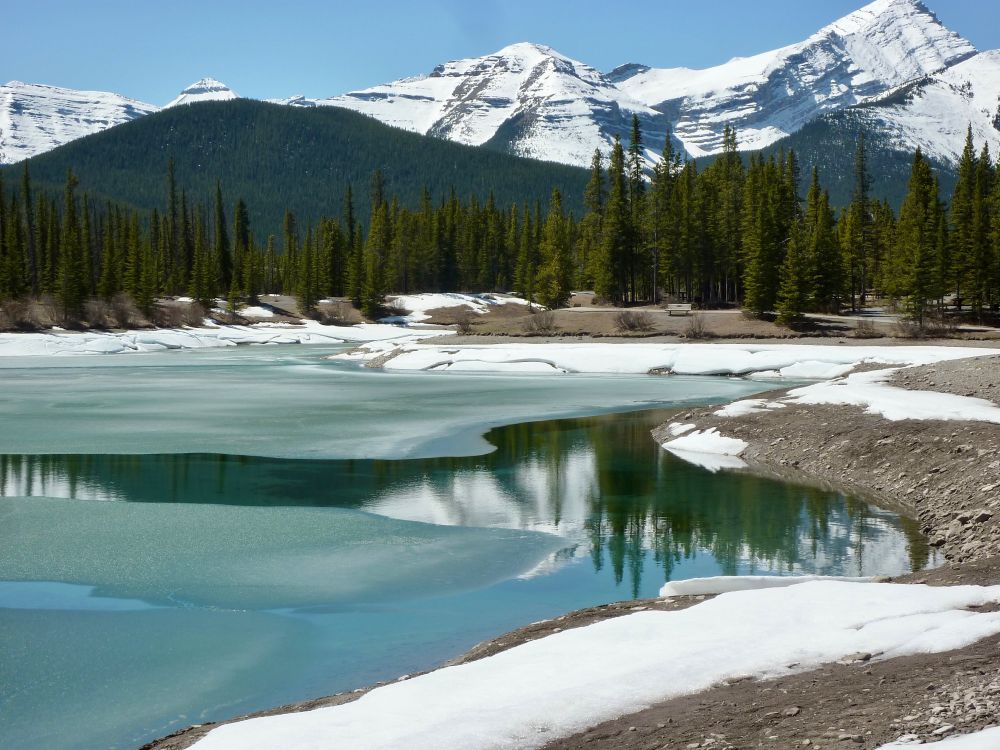 A partially frozen pond surrounded by snow covered mountains,
with a reflection in the open water.