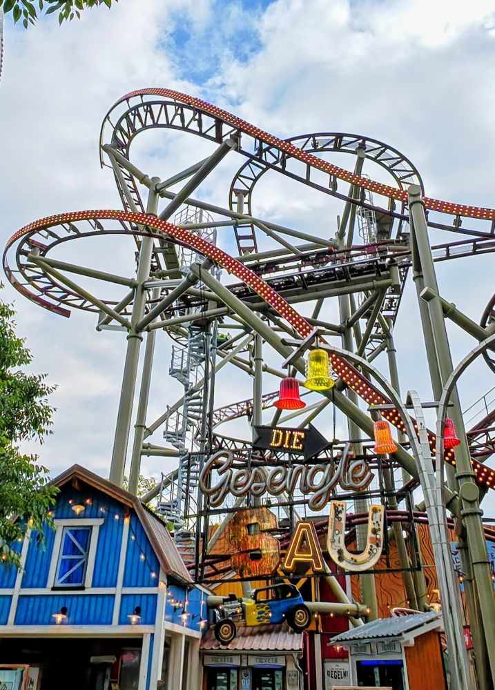 Eine wilde verschlungene Achterbahn vor bewölktem Himmel. Links unten davor steht ein kleines blaues Häuschen. An der Achterbahn steht in großen Lettern "Die gesengte SAU".