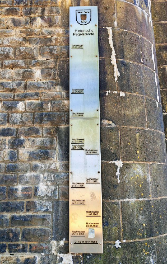 Eine historische Hochwassermarke an einer alten Steinmauer. Auf einer langen, schmalen Metalltafel sind verschiedene Wasserstände vergangener Hochwasserereignisse mit Datum eingetragen, zum Beispiel von 1855, 1880, 1920, 1946 und 1995. Die Tafel trägt die Überschrift „Historische Pegelstände“ und zeigt, wie hoch das Wasser in unterschiedlichen Jahren gestanden hat. Ganz oben ist das Stadtwappen von Wesel am Rhein zu sehen. 