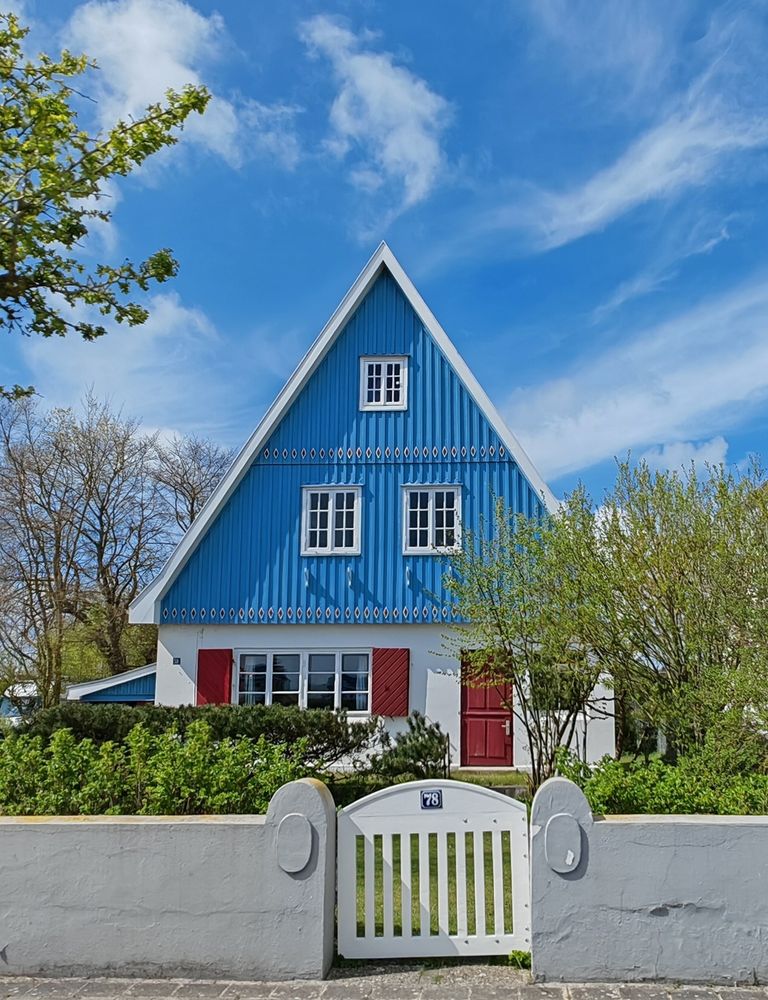 Ein blau gestrichenes Haus mit spitzem Giebeldach und weißen Fensterrahmen. Die Fassade ist oben vertikal mit blau gestrichenem Holz verkleidet und hat dekorative Muster in Rot, Weiß und Blau. Unten befinden sich rote Fensterläden und eine rote Eingangstür. Vor dem Haus steht ein weißer Gartenzaun mit Tor und der Hausnummer 78. Büsche und Bäume umrahmen das Gebäude, darüber ein blauer Himmel mit einigen weißen Wolken.
