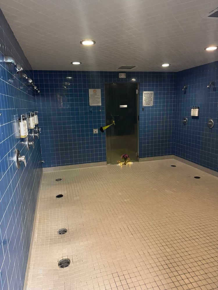 Open showers with 6 out of 11 shower heads in frame, the long closed sauna at the opposite side of the room. The blue tile walls and white tile floors telling stories of the many men who have frequented this spot. A small bouquet of flowers with electric tea candles are by the sauna door, setting a memorial for this soon to be closed relic. 