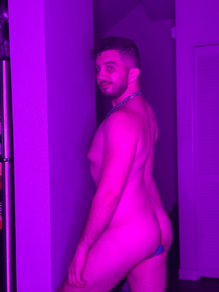 Nathan bathed in purple lighting, naked, looking over his left shoulder. The small spot of blue at the base of his ass, showing the plug he is holding in 