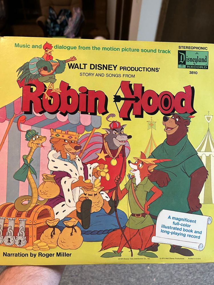 The album cover for the Walt Disney Productions 'Story and Songs from Robin Hood' (1973). The cover is a bright yellow and features a colorful illustration of characters from the animated film. The main characters, the fox Robin Hood and the bear Little John, stand on the right, smiling. On the left, a treasury scene shows Prince John (a lion) crying on a cushion surrounded by bags of gold, with Sir Hiss (a snake) looking on. The title 'Robin Hood' is in large red lettering across the top, with a rooster playing a lute perched above. A call-out on the lower right corner says, 'A magnificent full-color illustrated book and long-playing record.' The narration is credited to Roger Miller.